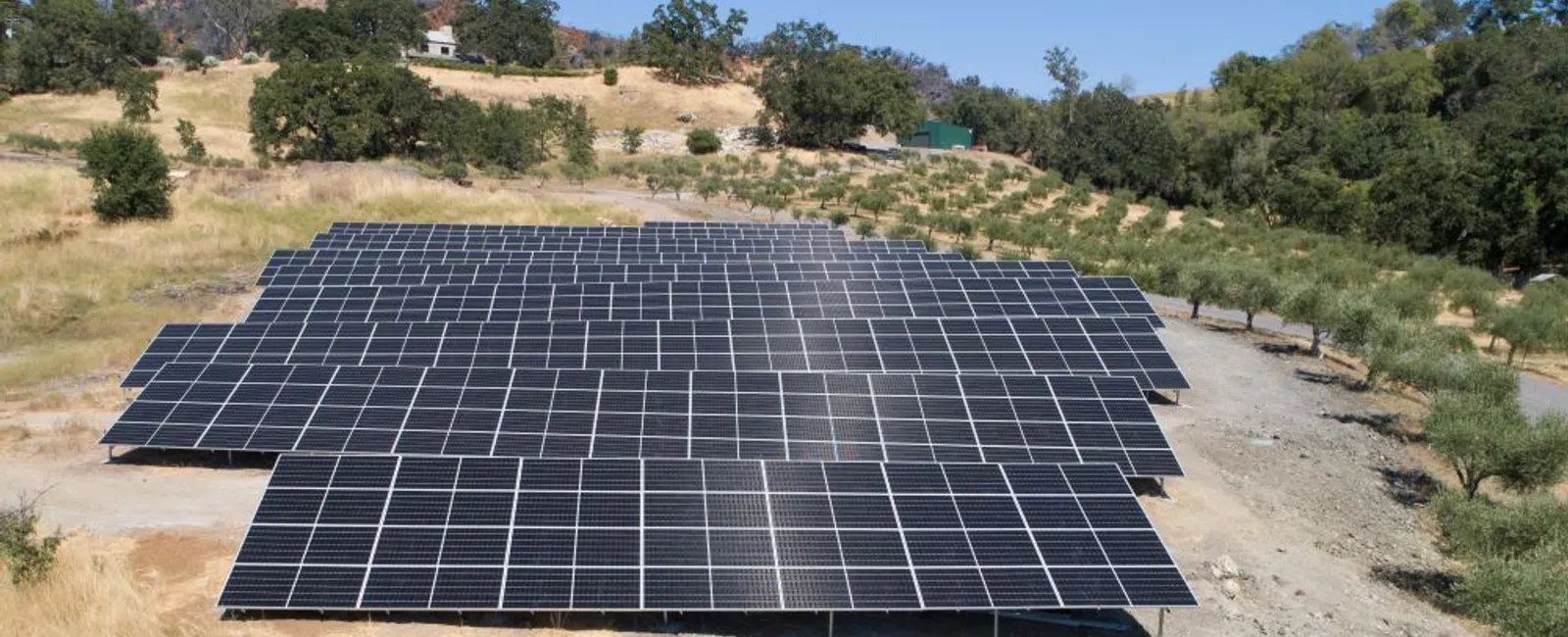 Aerial view of solar panels installed in a dry, grassy field surrounded by trees and hills.