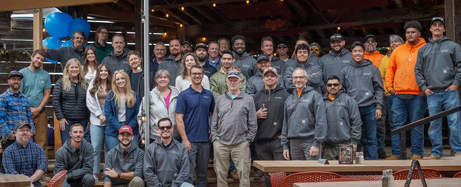 Group photo of a diverse team outdoors, smiling together under an awning at a lively event.
