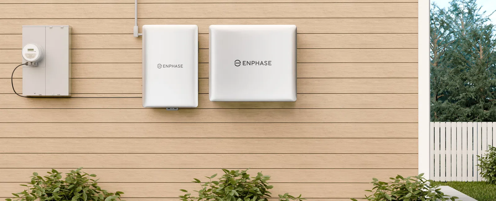 Enphase solar energy system components mounted on a light wood exterior wall above green shrubs in a backyard.