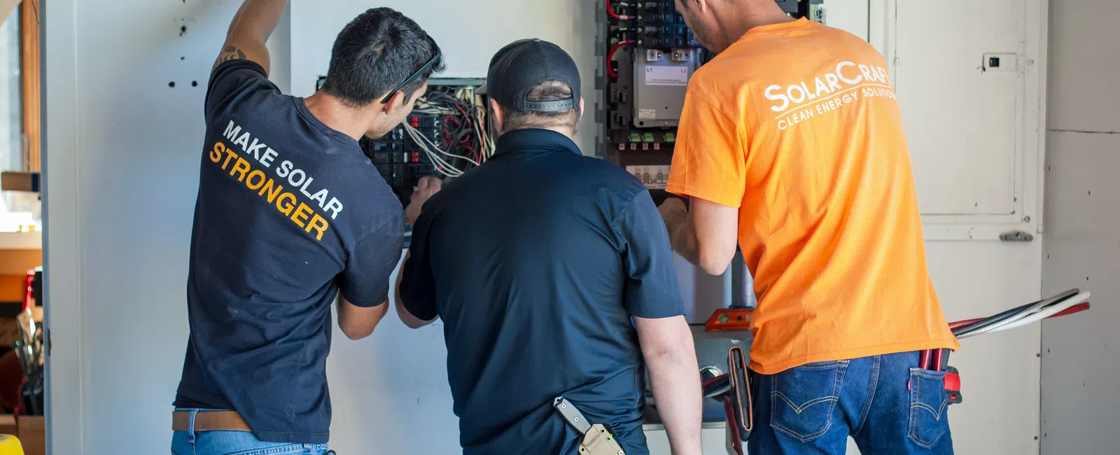 Three technicians working on electrical panels installing or repairing solar energy equipment indoors.