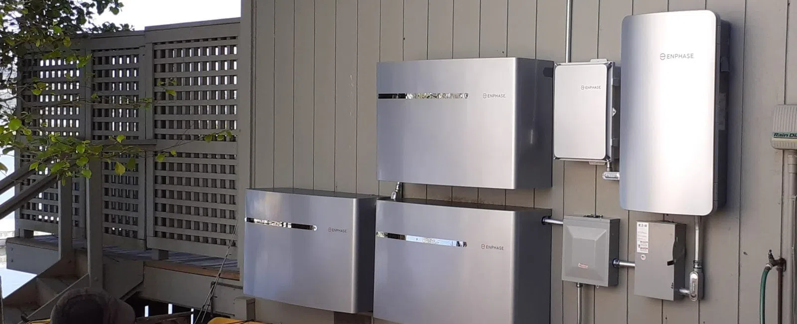 Home energy storage system with sleek silver batteries mounted on exterior wall above kayaks under tree shade.