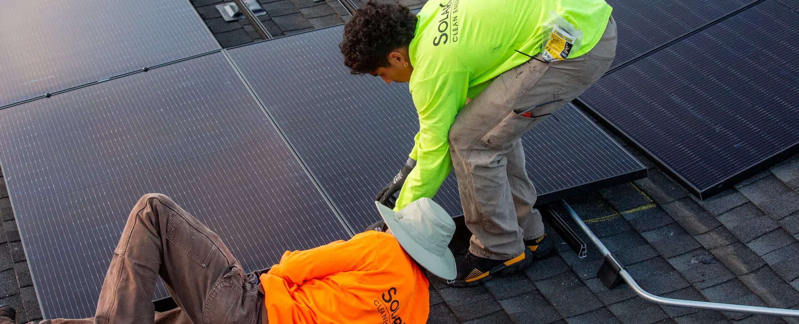 Two workers installing solar panels on a roof with safety gear and bright work shirts in daylight.