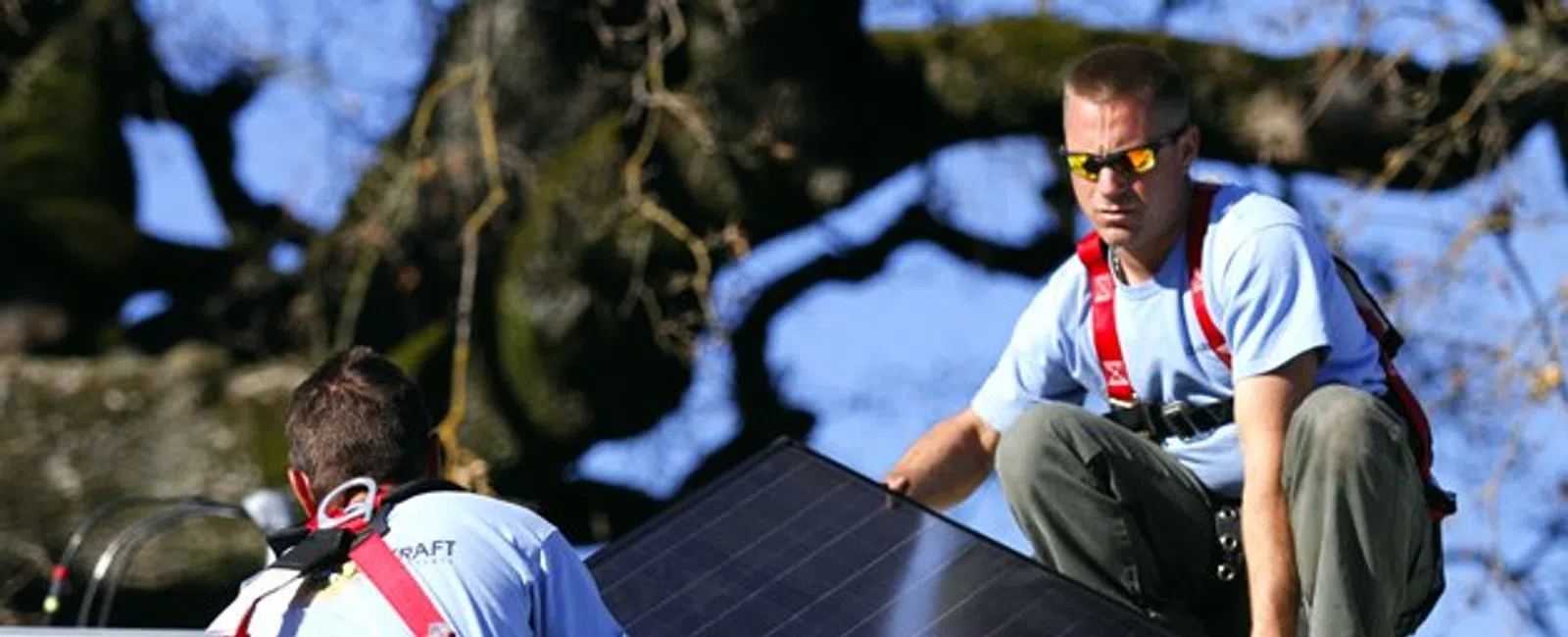 Two workers installing solar panels on a rooftop under clear blue sky with safety harnesses.
