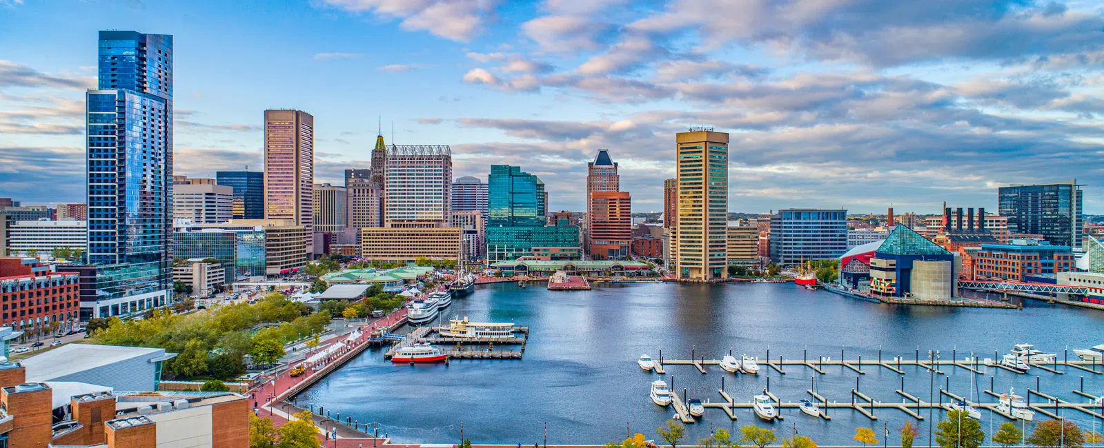 Baltimore Inner Harbor skyline with waterfront, boats, and modern buildings under partly cloudy sky.