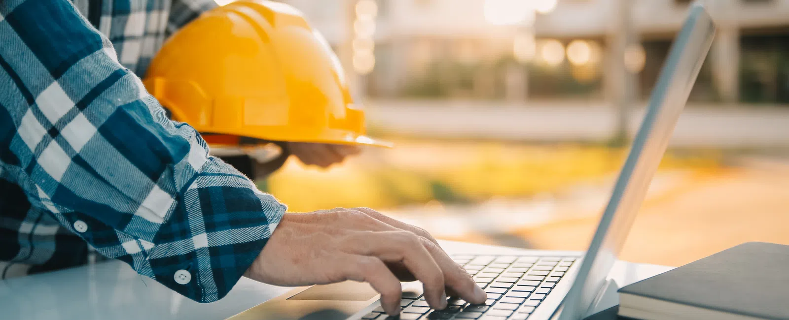 Construction worker wearing plaid shirt holding yellow hard hat while typing on laptop at desk.
