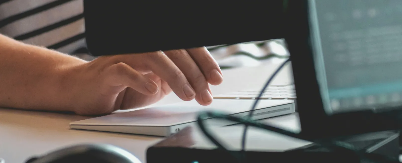 Person typing on a laptop keyboard with a mouse and monitors on a white desk in a dimly lit office.
