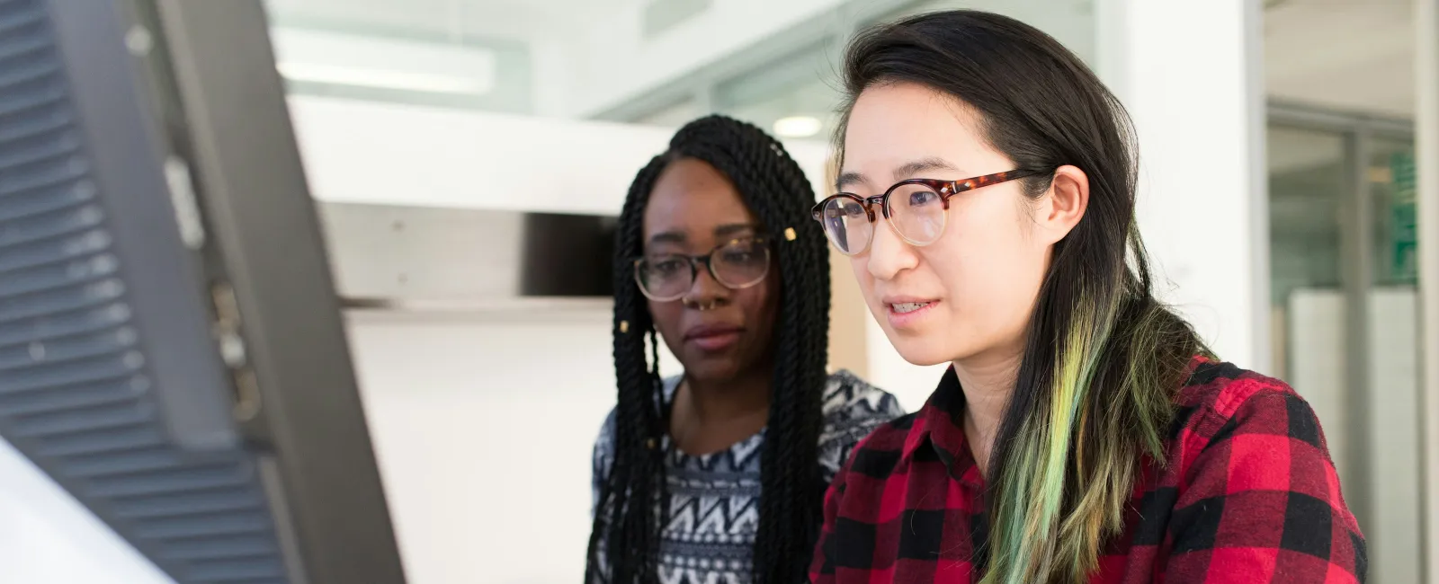 Two women collaborating and working together on a laptop in a modern office space with computer screens.