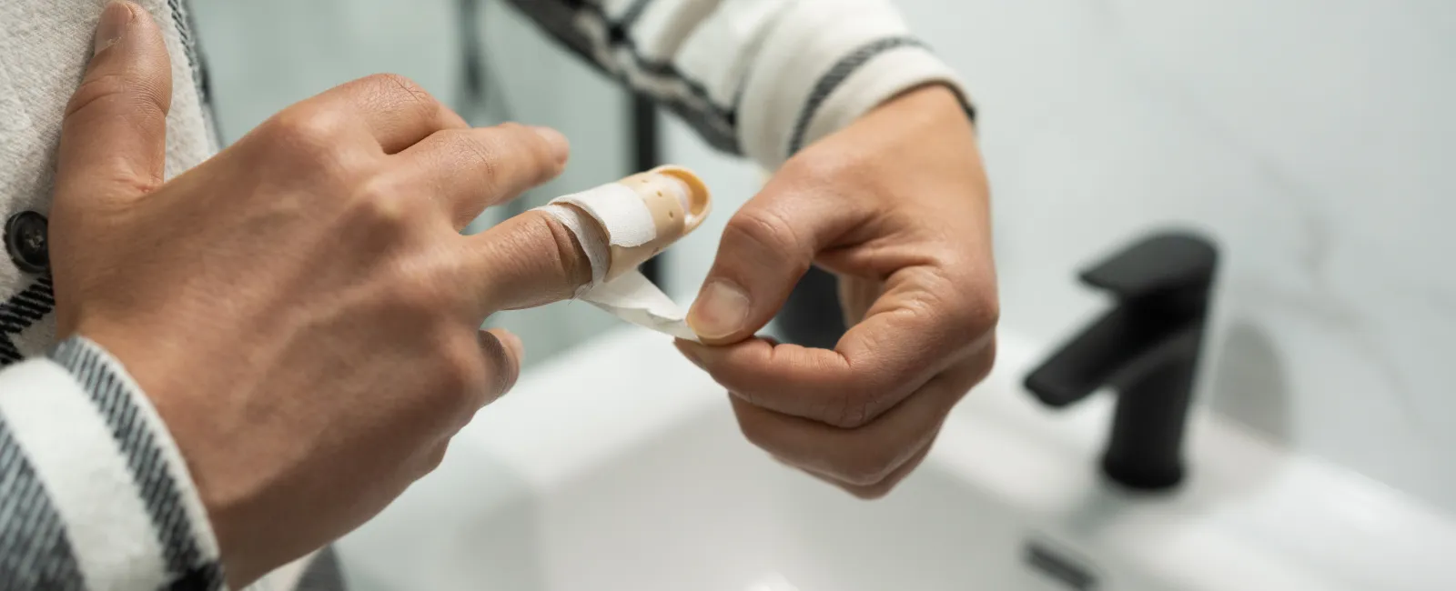 Person wrapping a bandage around an injured finger over a white sink with a black faucet.