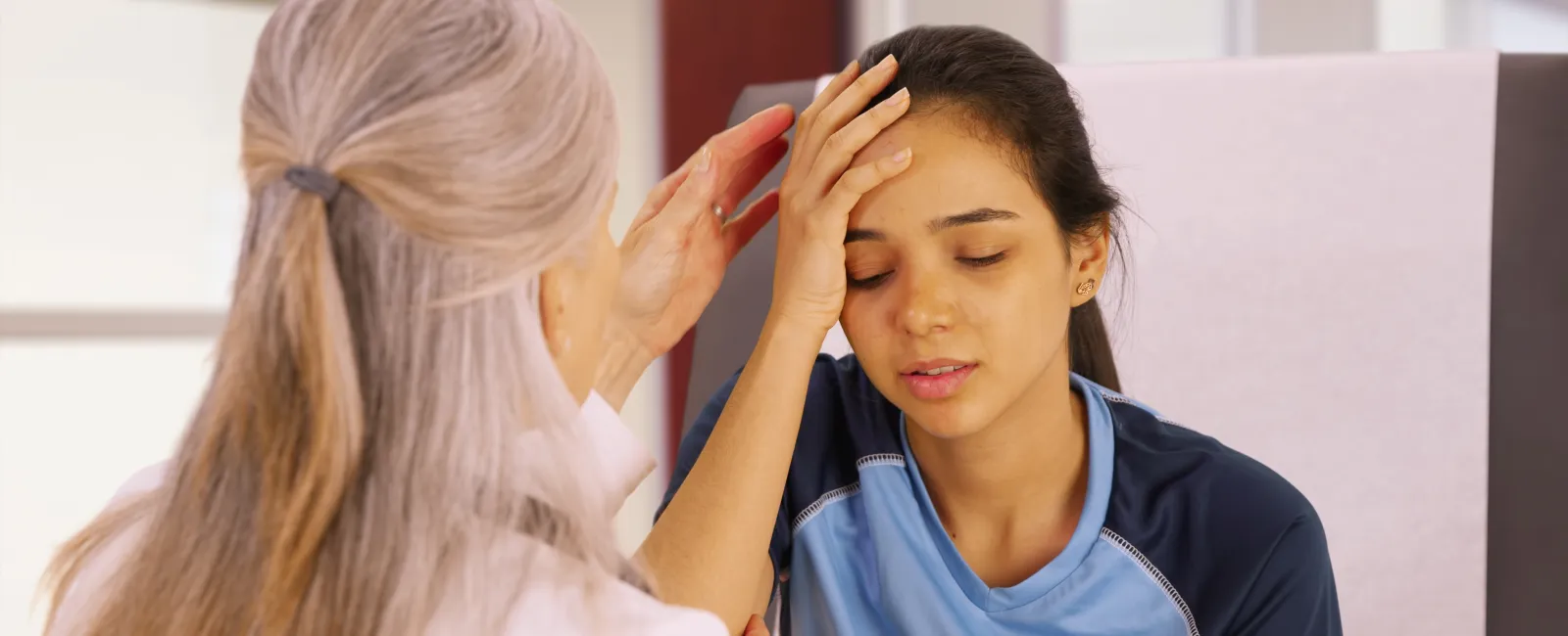 Young female athlete in blue jersey with headache being comforted by older woman in a medical setting
