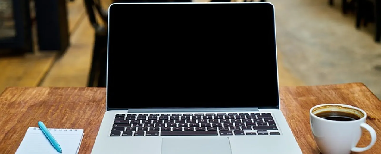 Laptop with blank screen, notebook with pen, and cup of coffee on wooden table in a cafe setting.