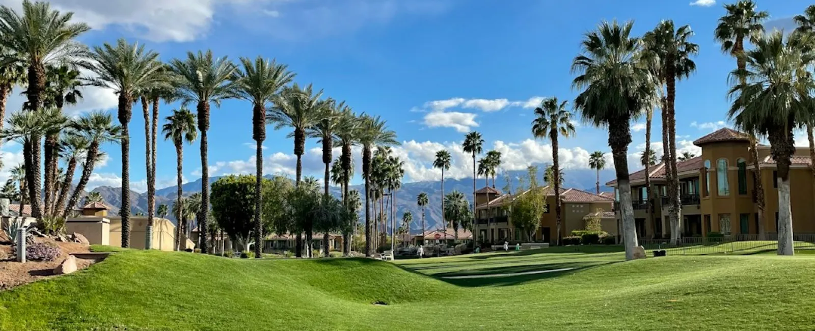 Green golf course with palm trees and houses reflected in a calm pond under a partly cloudy blue sky.
