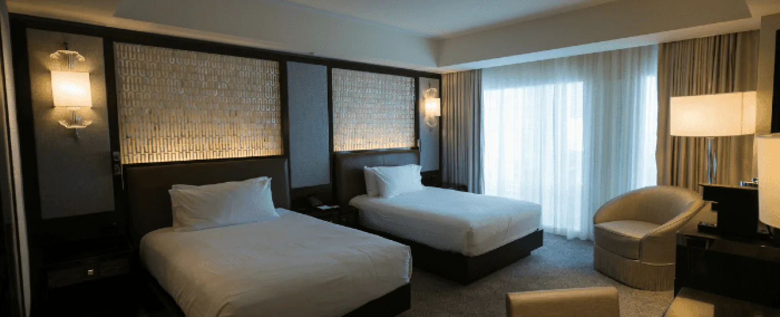Modern hotel room with two beds, upholstered chair, desk, soft lighting, and large window with curtains.
