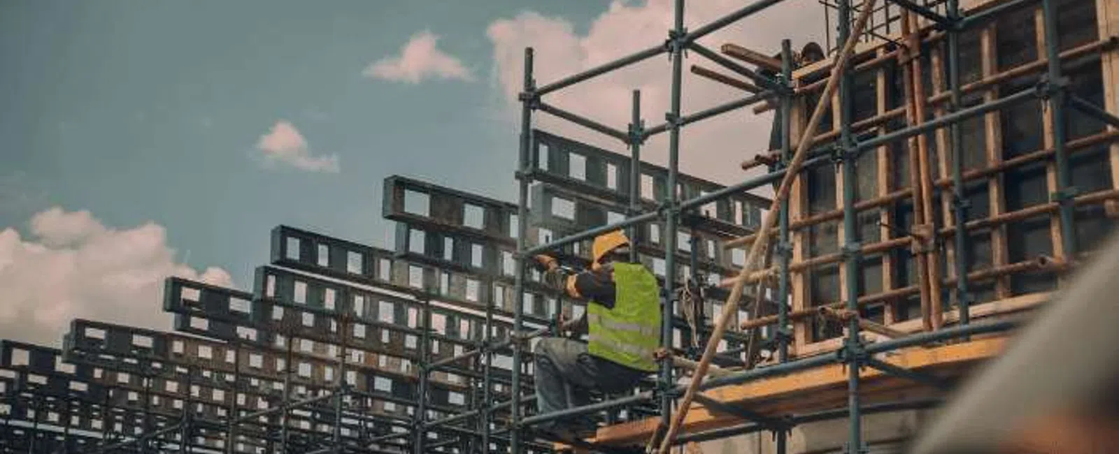 Construction worker in safety vest on scaffolding at an urban building site under blue sky with clouds.