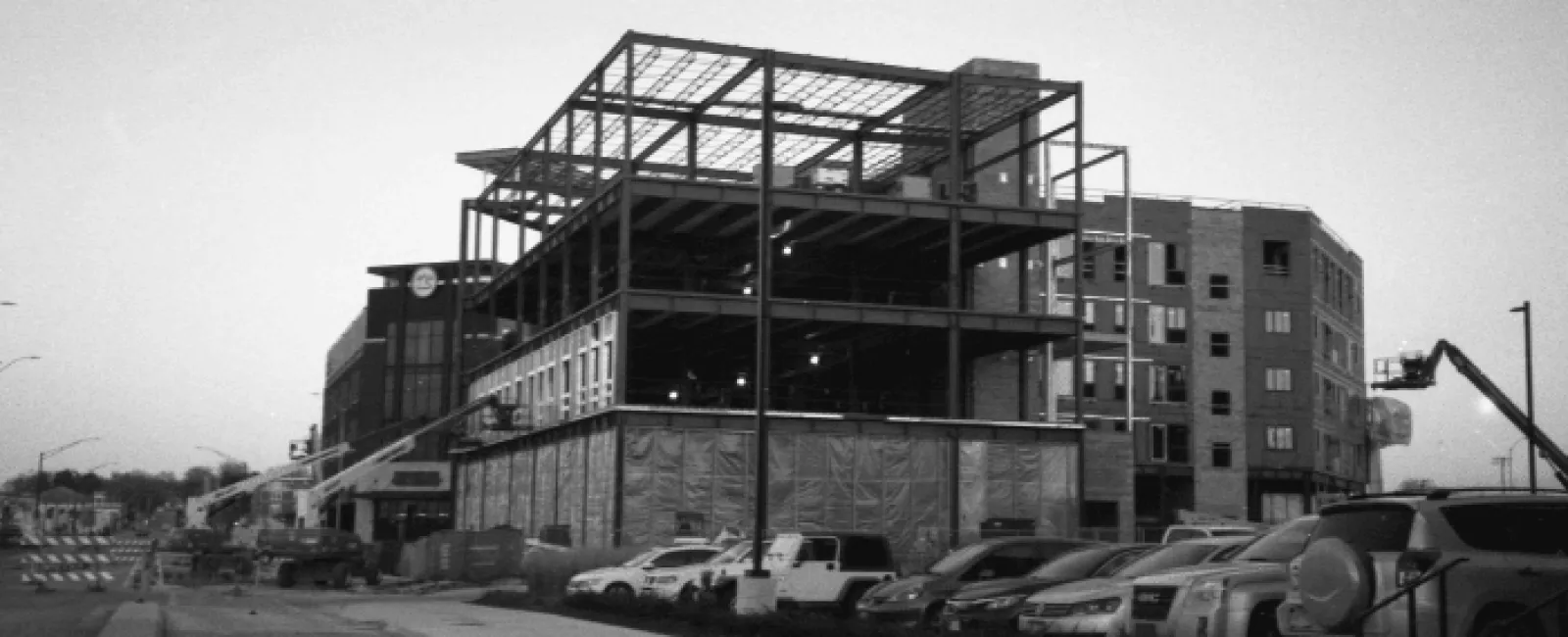 Black and white photo of a multi-story building under construction with parked cars along the street.
