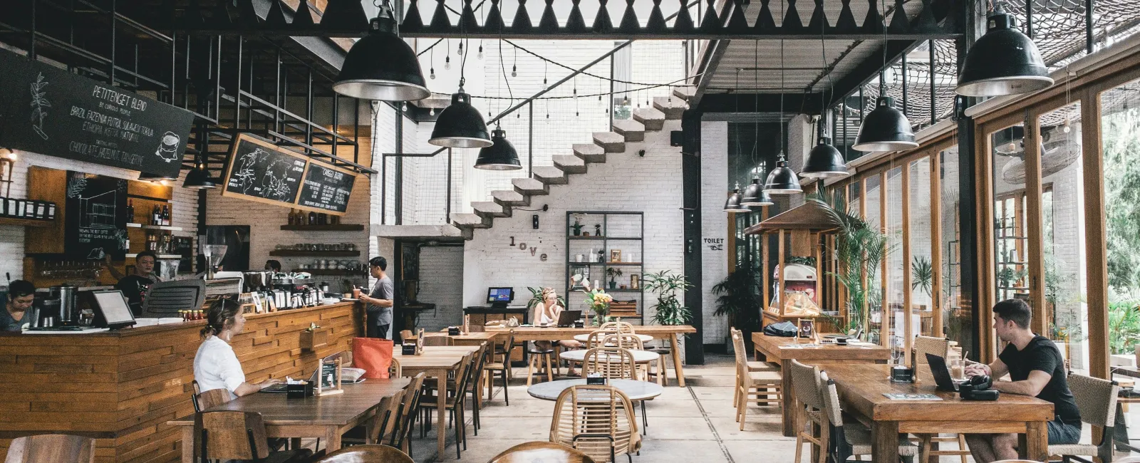 Spacious modern cafe interior with wooden furniture, industrial lighting, and large windows letting in natural light.