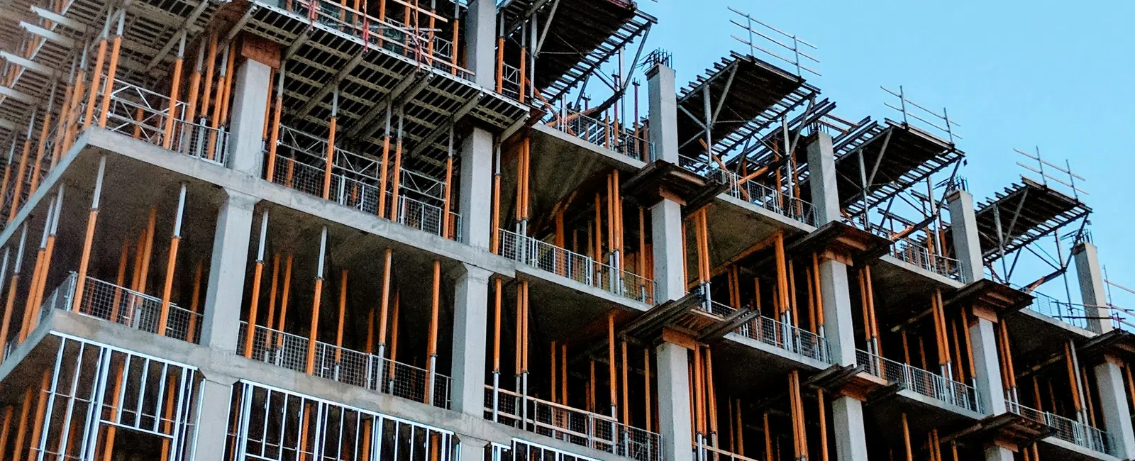 Under-construction multi-story concrete building with scaffolding and steel reinforcements against a clear blue sky.