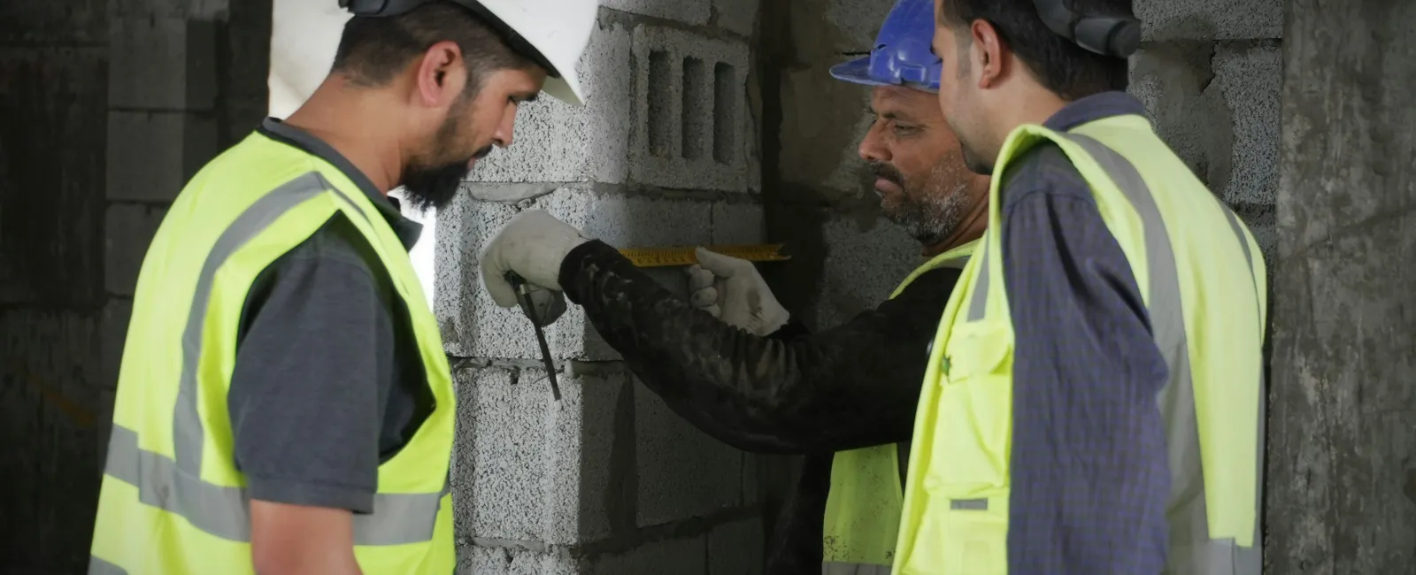 Three construction workers in helmets and reflective vests measuring a concrete block wall inside a building