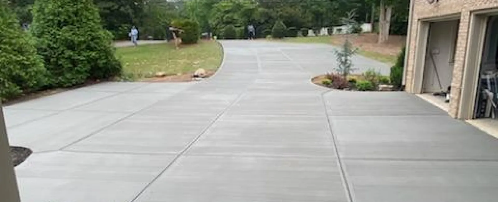 Wide concrete driveway next to brick house with greenery and trees in the background on a cloudy day