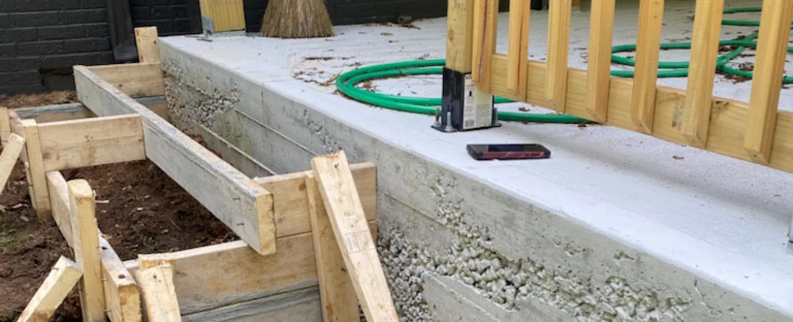 Wooden framing and freshly poured concrete foundation for outdoor stairs next to a porch with broom and hose.