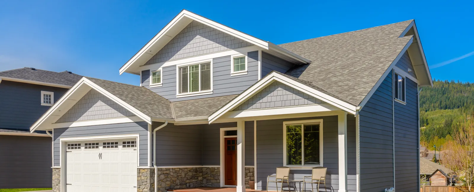 Modern two-story gray house with stone accents, front porch, garage, and green lawn under clear blue sky.
