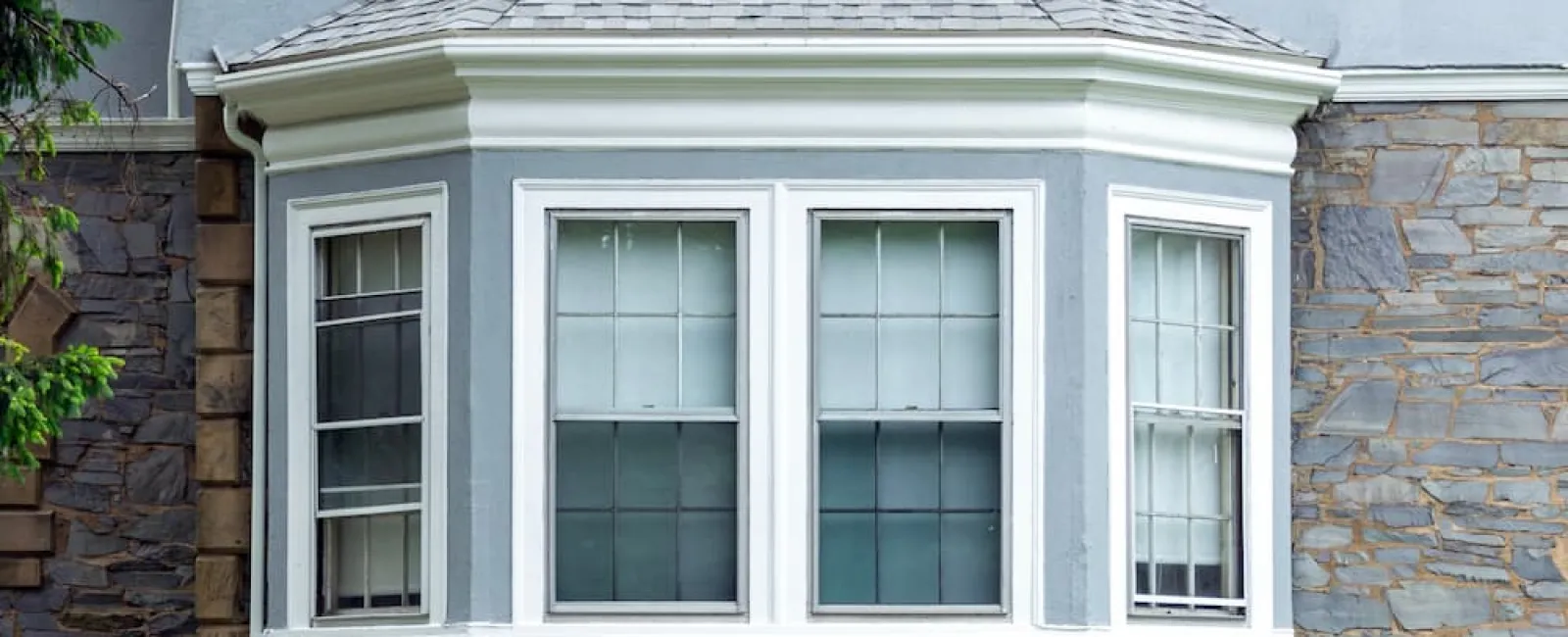 Exterior view of a grey stone and stucco house featuring white historic home windows and a classic bay window design.