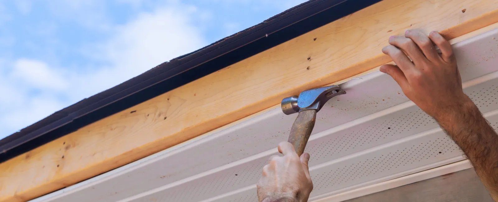 Close-up of a person using a hammer to install wooden fascia board under a roof against blue sky.