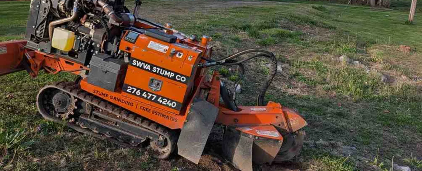 Orange stump grinding machine operating on grassy residential lawn near houses and trees on a clear day.