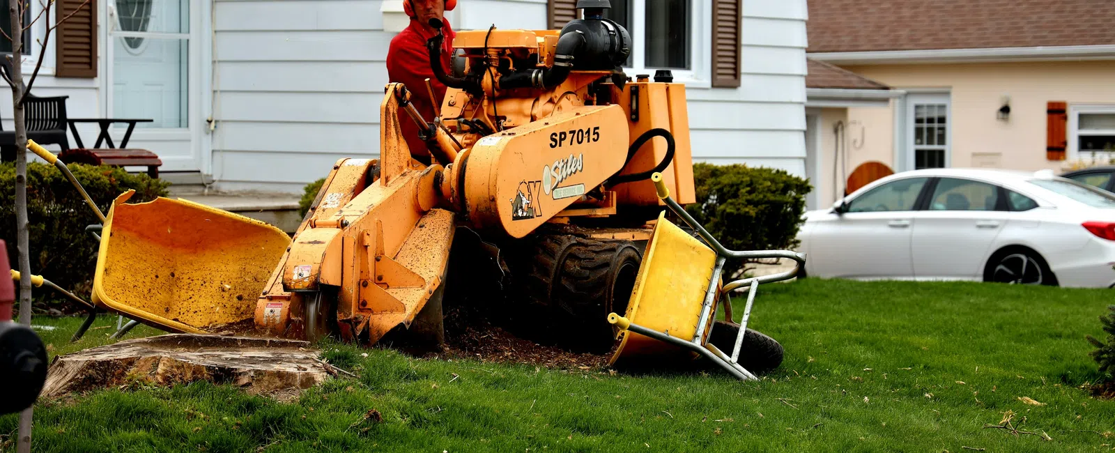 Worker operating stump grinder machine removing tree stump in residential front yard with houses in background.