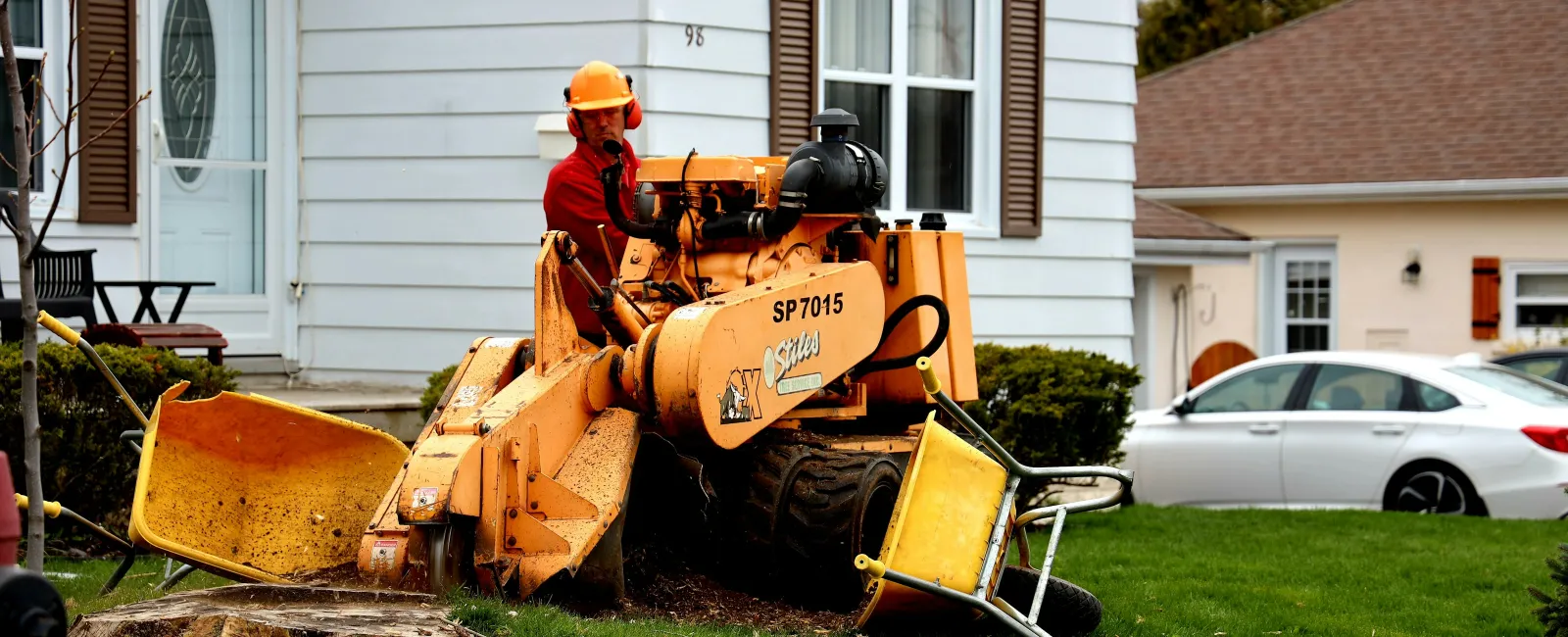 Worker using stump grinder machine to remove tree stump in residential lawn with houses and car in background
