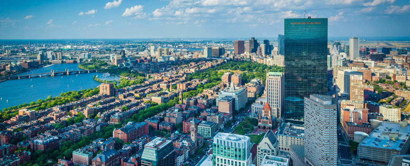 Aerial view of Boston city skyline with river, skyscrapers, and green parks under a blue sky with clouds.