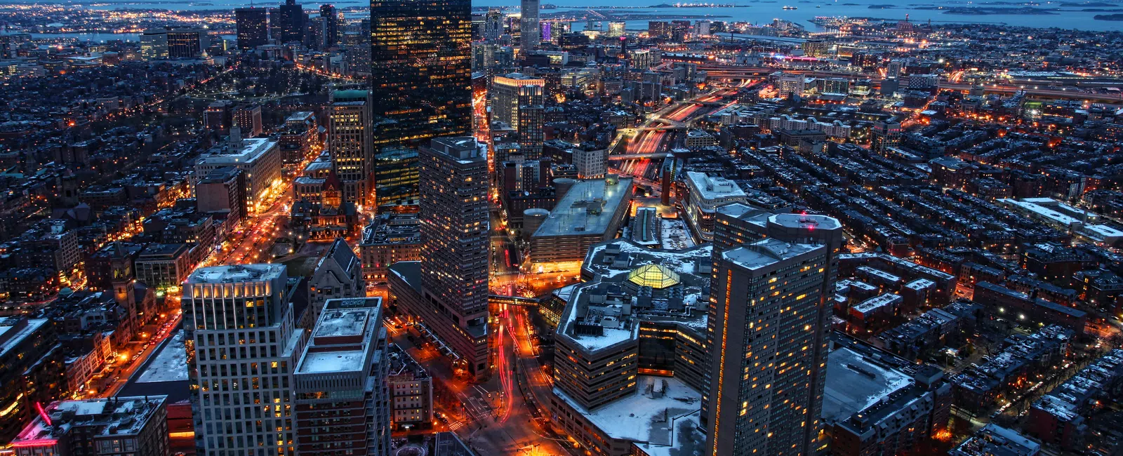 Aerial view of a city skyline at dusk with illuminated buildings, streets, and snowy rooftops.