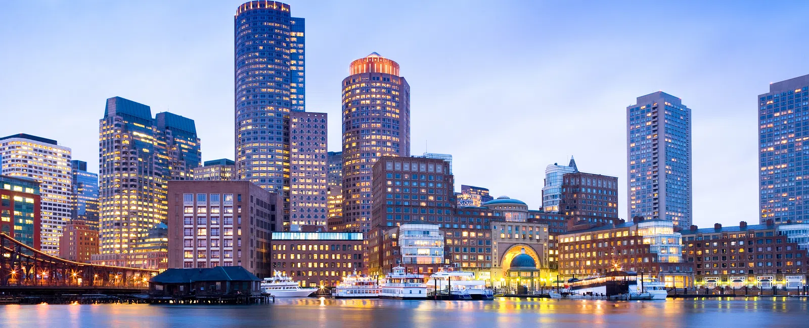 Boston waterfront skyline at dusk with illuminated buildings and calm reflective water under a clear sky