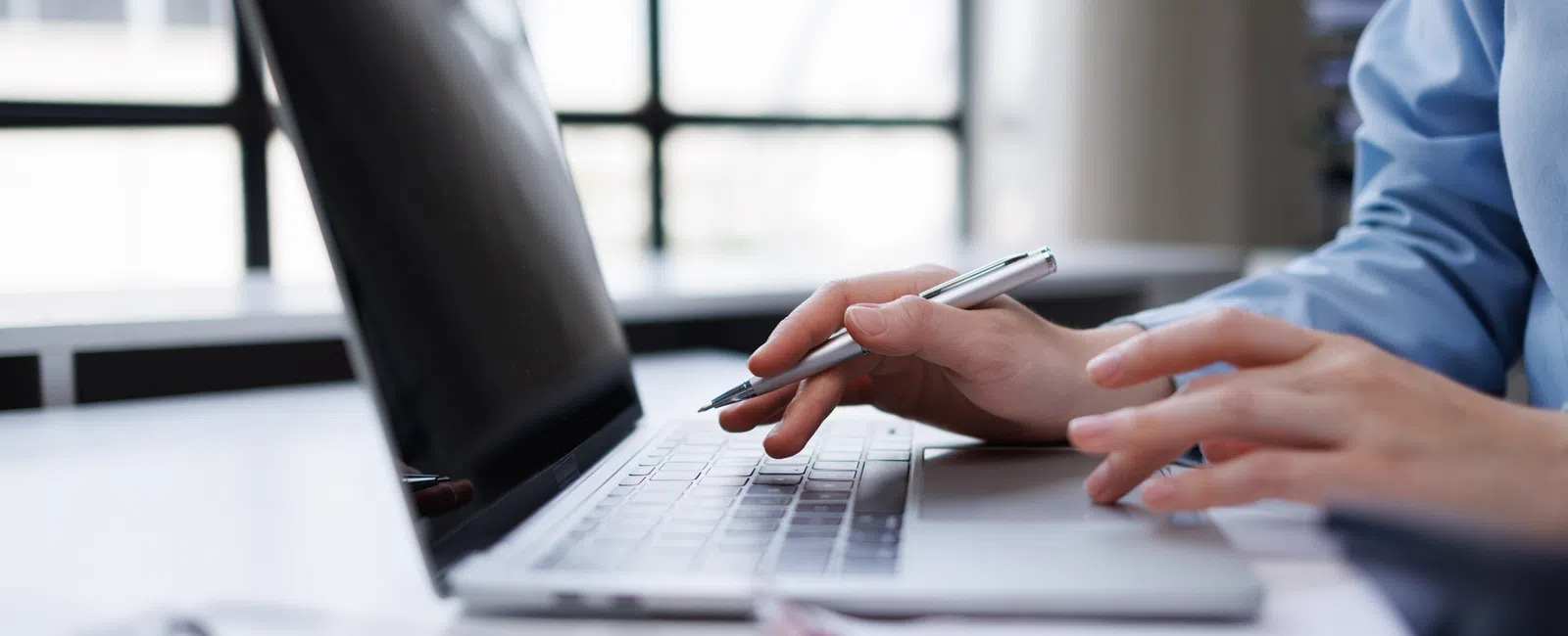 Close-up of hands using a laptop keyboard and holding a pen in a bright modern office setting.