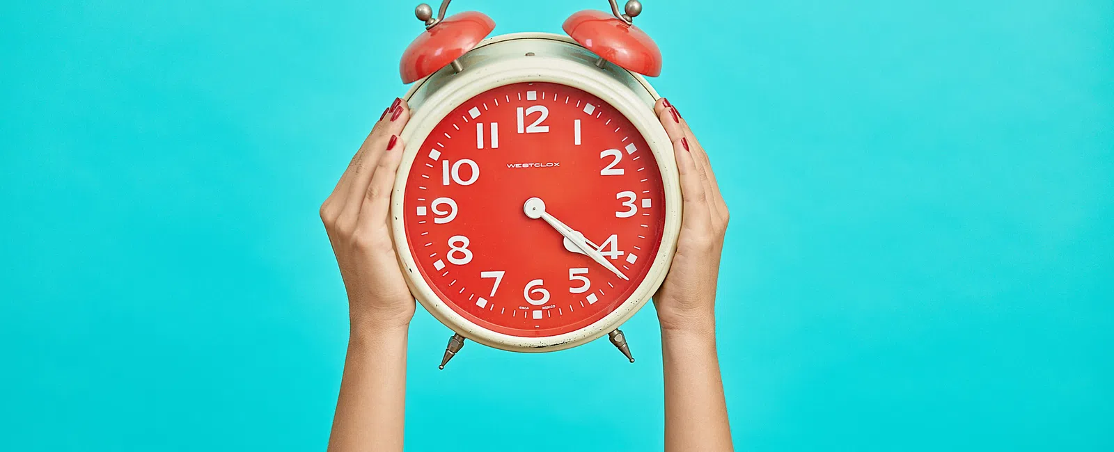 Hands holding a large red alarm clock against a bright turquoise background showing 4:20 time.