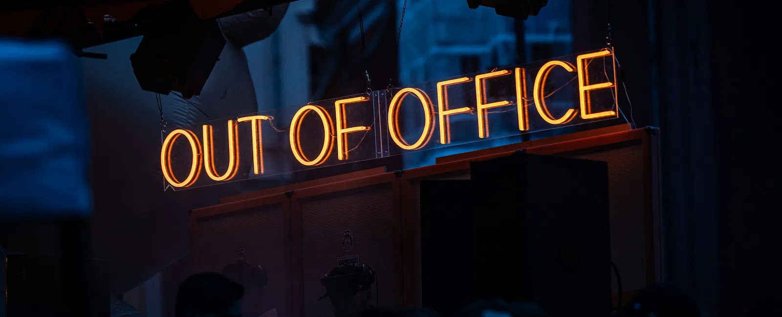 Glowing orange neon sign reading OUT OF OFFICE hanging indoors with people in dark foreground