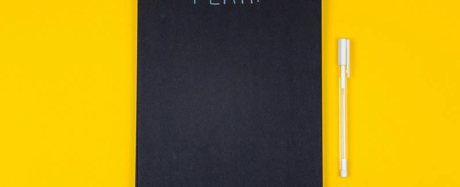 Black spiral notebook with PLAN written on cover and a white pen on a yellow background.