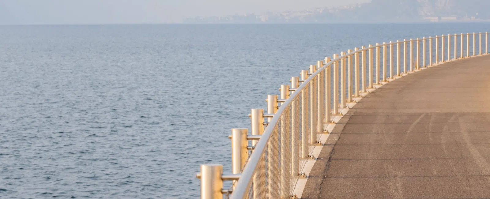Curved waterfront pathway with metal railing beside calm sea under hazy sky and distant mountains.