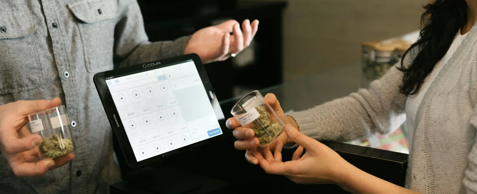 Two people exchanging cannabis samples at a counter with a digital point-of-sTwo people exchanging small containers of cannabis at a cannabis dispensary with digital point-of-sale tablet up per cannabis IT compliance.ale tablet and glass containers.