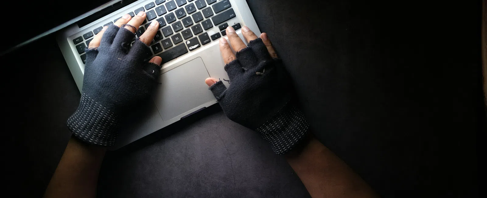 Hands wearing fingerless gloves typing on a laptop keyboard against a dark background.