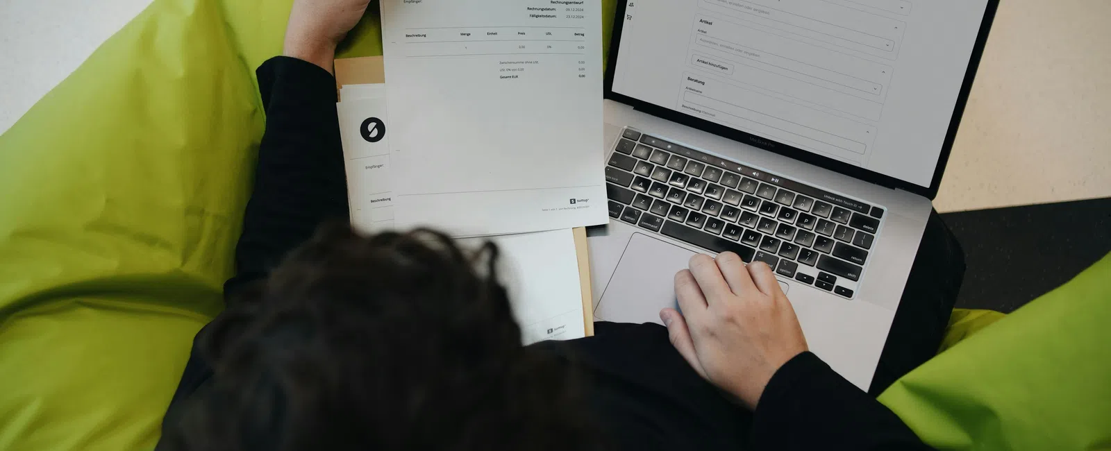 Person reviewing documents and entering information on a laptop while sitting on a green bean bag chair.