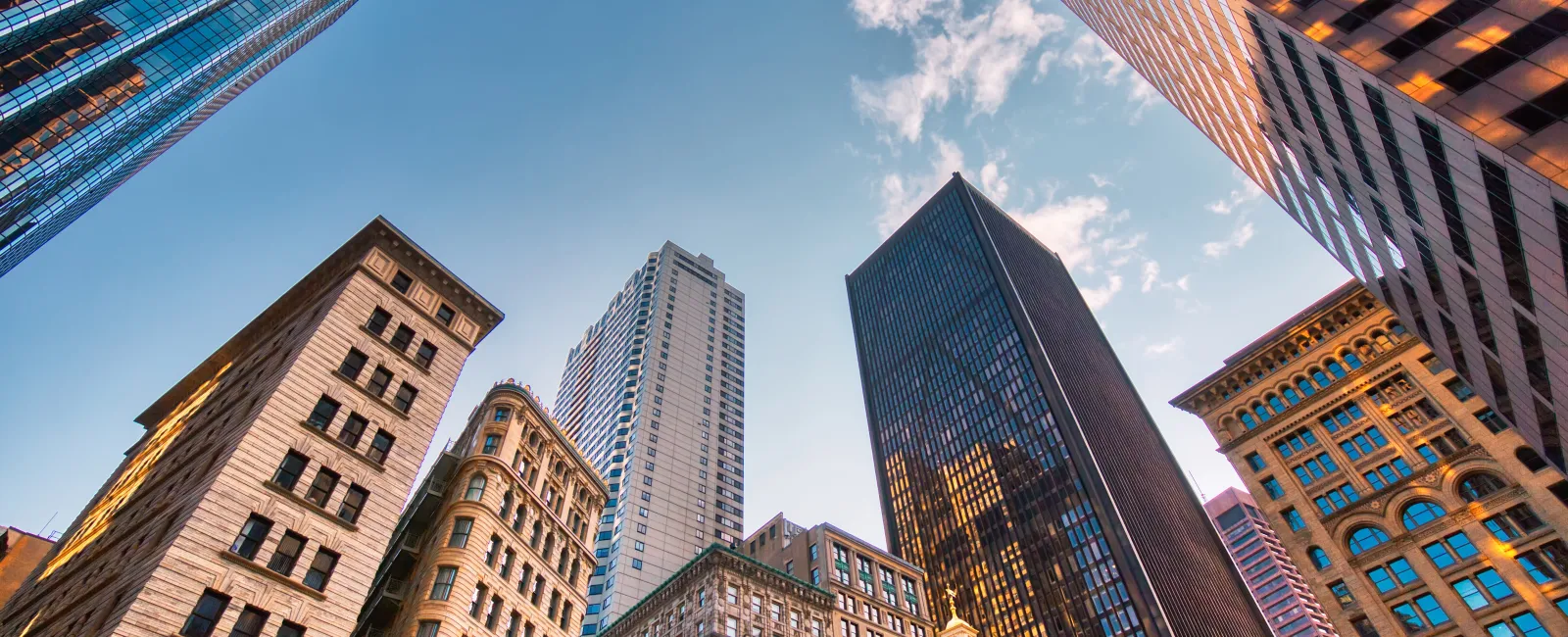 Upward view of diverse skyscrapers and historic buildings under a partly cloudy blue sky.