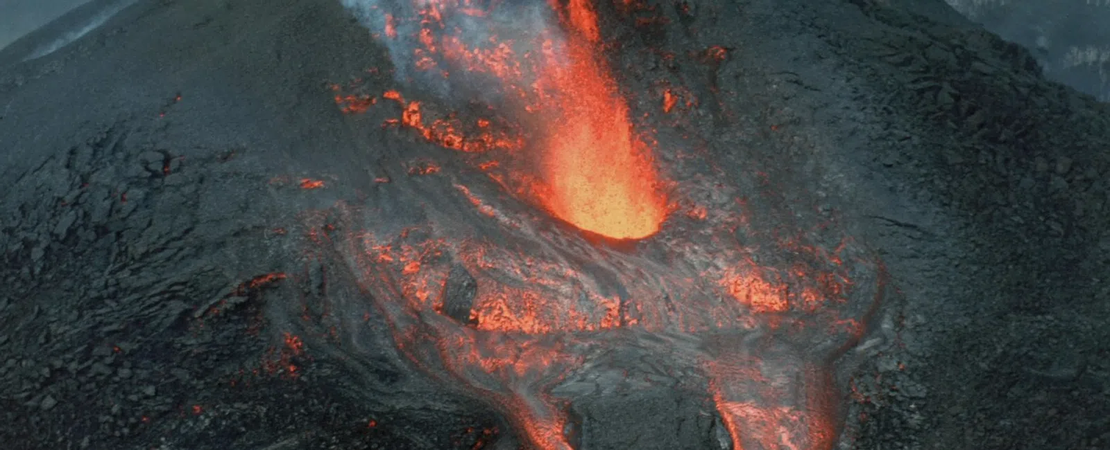 Active volcano erupting with flowing red hot lava and thick white smoke against dark volcanic rock.