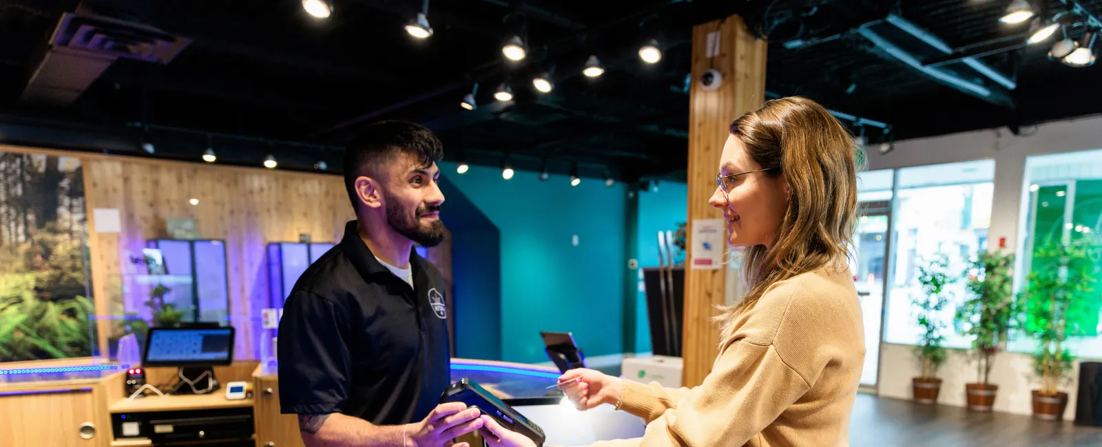 Customer uses contactless payment at store counter assisted by smiling male employee in casual retail setting.