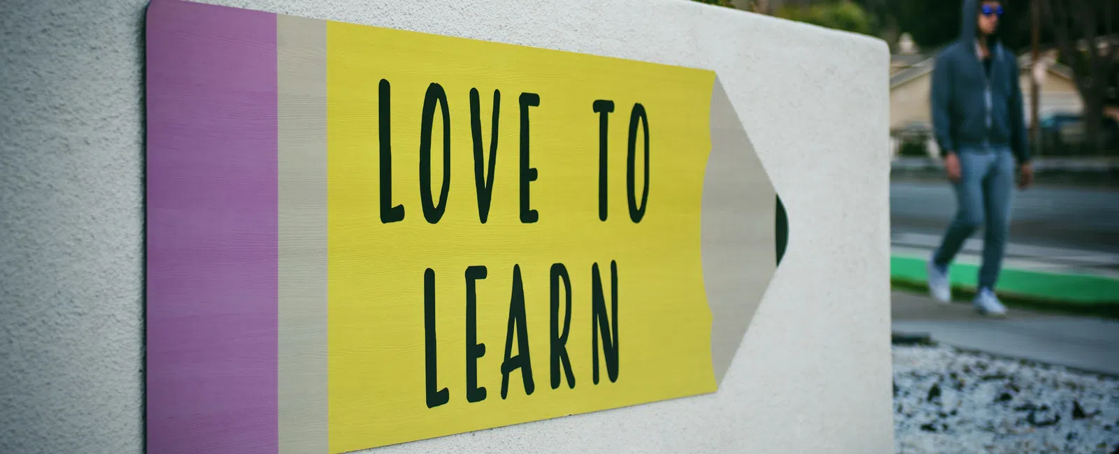 Large pencil-shaped sign on white wall with message Love to Learn in black text outdoors