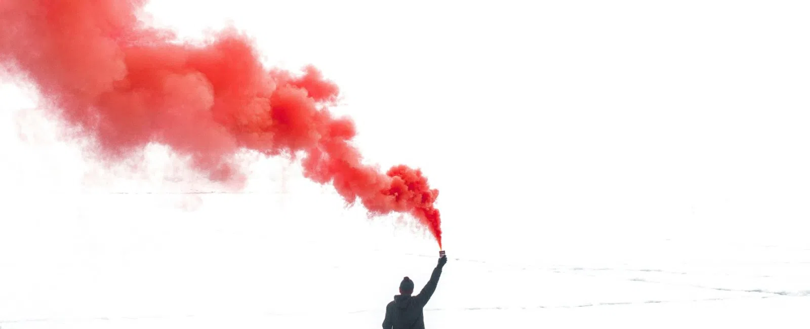 Person in black stands on icy terrain holding a red smoke flare against a bright white sky.