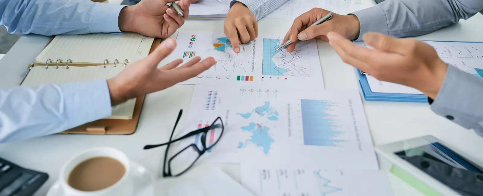 People collaborating over financial charts and data sheets with coffee and glasses on a white office table.