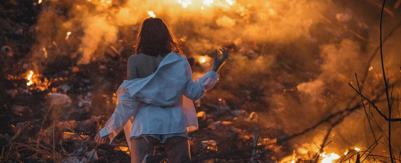 Woman in white shirt stands facing a large fire burning in a dark, outdoor environment at night.