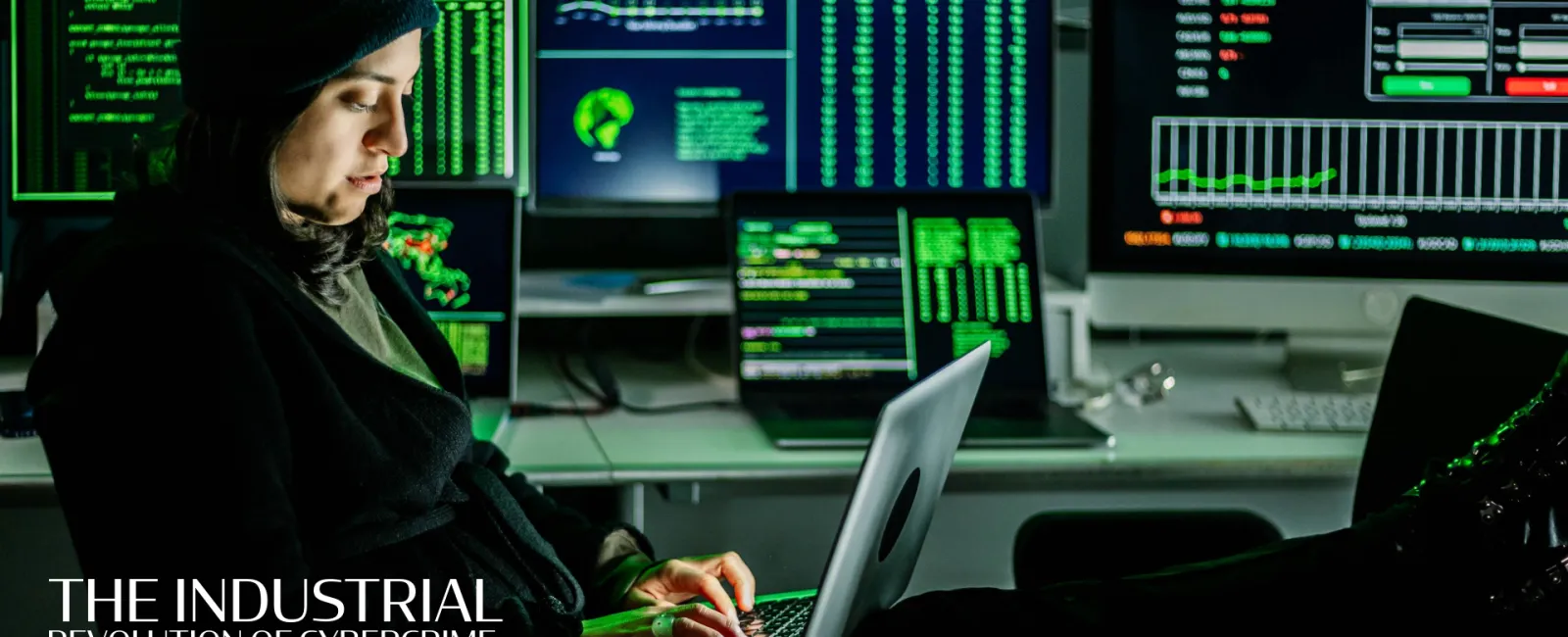 Person in dark clothing working on laptop surrounded by multiple monitors displaying code and data.