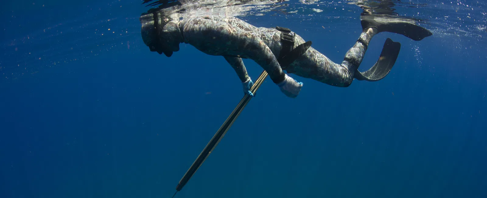 Freediver in camouflage wetsuit holding speargun underwater in clear deep blue ocean with fins.