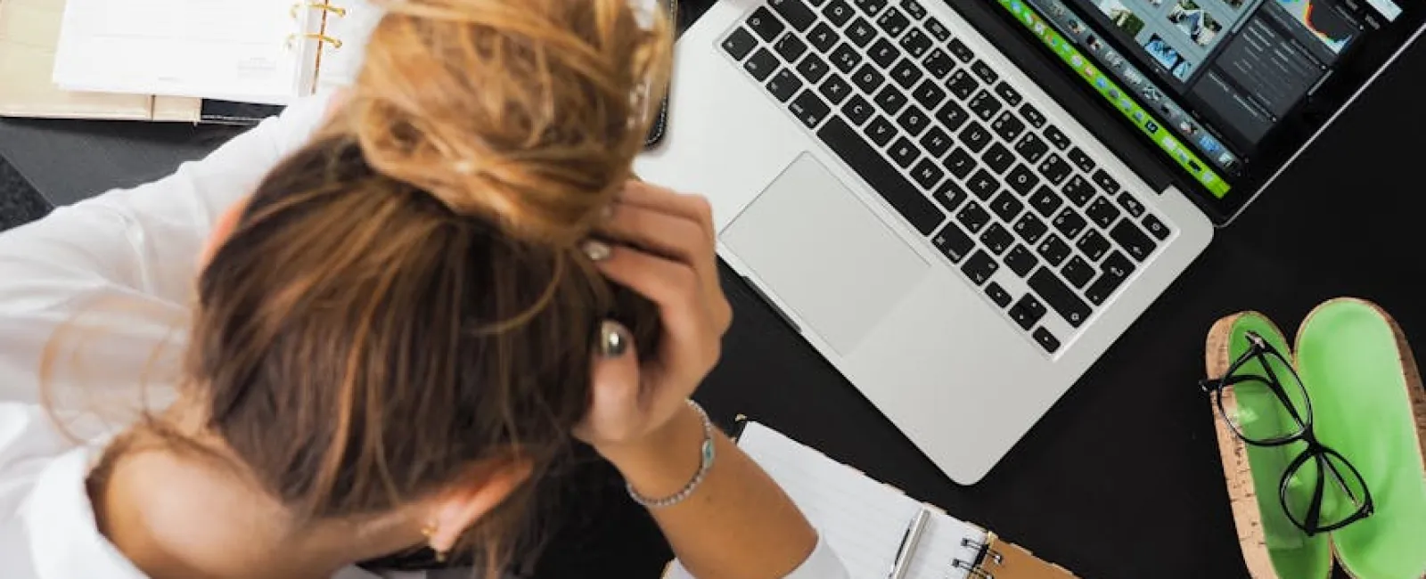 Overhead view of a stressed woman working at a desk with laptop, notebooks, and smartphone, holding her head.